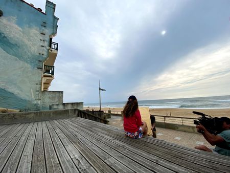 Big wave surfer Justine Dupont sits on steps on the boardwalk next to the beach. DP Alfredo de Juan films from below and AC Florian Morel holds the light reflector.  (National Geographic/Gene Gallerano)