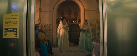 The elevator doors at Naples National Museum of Archaeology have opened on a gathering at the shrine of the goddess Isis at Julia Felix's estate.  Julia Felix, played by Lubna Azabal, leads prayers and offerings at the altar, assisted by Amalthea, played by Buket Komur, who holds a platter of dates and honey as offerings to the goddess. (National Geographic)