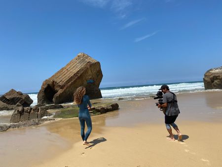 Big wave surfer Justine Dupont, wearing her wetsuit, walks on the beach with her feet in the water. DP Alfredo de Juan follows her, filming.  (National Geographic/Gene Gallerano)
