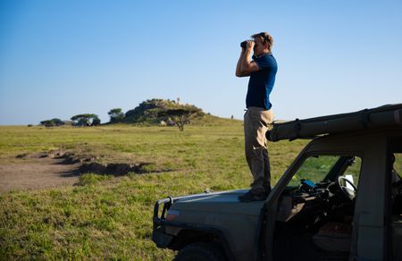Bertie Gregory stands on the front bonnet of a safari Jeep looking for animals through binoculars. (credit: National Geographic/Jigar Ganatra)