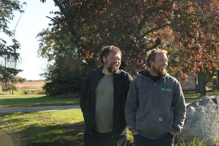 Charles Pol and Ben Reinhold laugh in front of the large rock they are planning to move to the farm's new garden. (National Geographic)