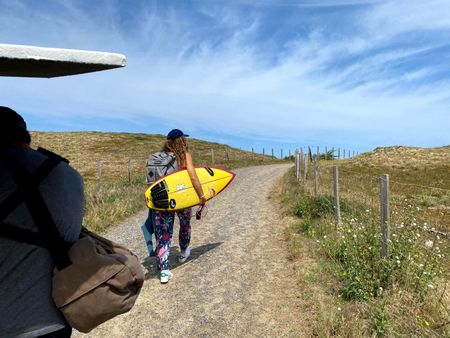 Big wave surfer Justine Dupont walks on a trail that leads to the beach with her board under an arm. Crew is following her.  (National Geographic/Gene Gallerano)