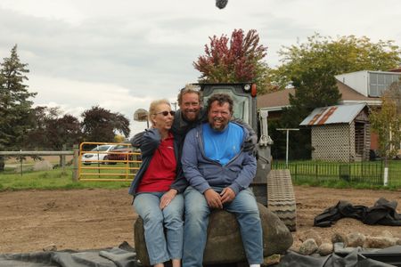 Diane Pol, Ben Reinhold, and Charles Pol smile while sitting on a rock at the Pol family farm's new pond and waterfall. (National Geographic)