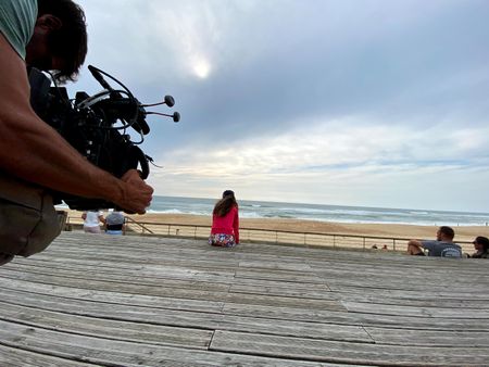 Big wave surfer Justine Dupont sits on steps on the boardwalk next to the beach. DP Alfredo de Juan films from behind.  (National Geographic/Gene Gallerano)