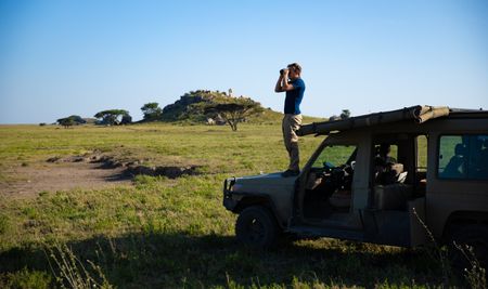 Bertie Gregory stands on the front bonnet of a safari Jeep looking for animals through binoculars. (credit: National Geographic/Jigar Ganatra)