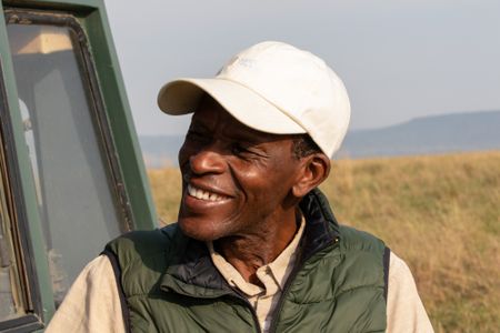 Lead film driver Sammy Munene smiles while waiting by the vehicle he drives, taking a break as Summer's Pride relaxes nearby. (National Geographic/Joe Treddenick)