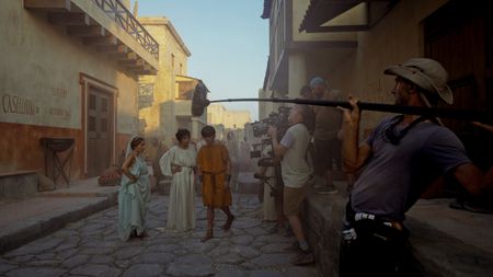 Behind-the-scenes showing Buket Komur (Amalthea), Lubna Azabal (Julia Felix), and Frankie Treadaway (Avianius) filming a recreation scene with Director of Photography Jim Jolliffe operating the main camera and support crew behind him. (National Geographic/Matthew Muscat Drago)