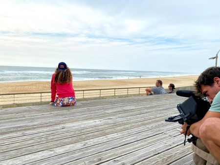 Big wave surfer Justine Dupont sits on steps on the boardwalk next to the beach. DP Alfredo de Juan films from behind.  (National Geographic/Gene Gallerano)