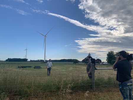A crew member films Ben Reinhold and Charles Pol holding a tape measure in the field, while barn cat Mr. Hyde watches on from on top of a post. (National Geographic)