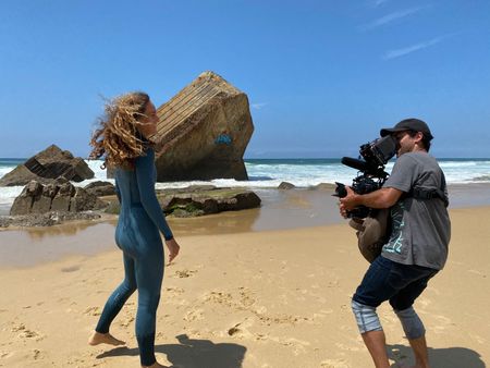 Big wave surfer Justine Dupont, wearing her wetsuit, walks on the beach with her feet in the water. DP Alfredo de Juan follows her, filming.  (National Geographic/Gene Gallerano)