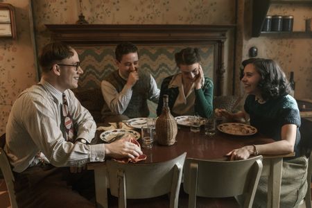 A SMALL LIGHT - Jan, Kuno, Hendrika and Miep laugh over breakfast in A SMALL LIGHT. (From left: Joe Cole as Jan Gies, Preston Nyman as Kuno, Gabriella Farr as Hendrika, and Bel Powley as Miep Gies). (Credit: National Geographic for Disney/Dusan Martincek)