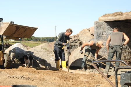Dax, Ben's dog, stands by Ben Reinhold as he uses a mallet to push a pole into the ground while Seth Doble and Scott Brady level out the wet concrete they are using for the sheep hut's flooring. (National Geographic)