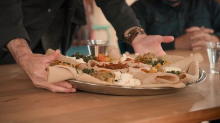 An injera platter being placed on the table by host Stanley Tucci where another participant of Cooking Without Borders is seated.  (credit: National Geographic)