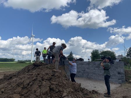 Carter Korthals, Kyle Knoche, Scott Brady, Charles Pol, Tobias Edgett, and Andrew Hutton stack cement blocks for the Pol family farm's sheep hut wall, while a crew member films them. (National Geographic)